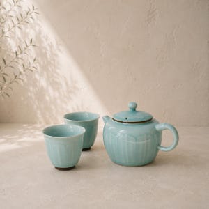 Op de afbeelding: Een lichtblauwe keramische theeset, bestaande uit een theepot met deksel en twee bijpassende kopjes. De theepot en kopjes hebben een geribbeld ontwerp. De set wordt gepresenteerd op een neutrale ondergrond, met zachte verlichting.