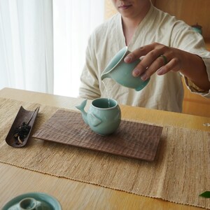 May include: A person pouring water from a light teal ceramic pitcher into a matching whale-shaped tea cup. The cup sits on a carved wooden tray, with a bamboo tray of tea leaves to the left. The scene is set on a woven table runner.