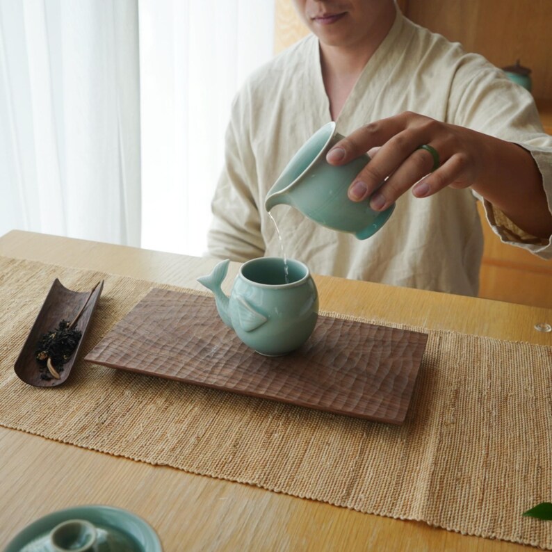 May include: A person pouring water from a light teal ceramic pitcher into a matching whale-shaped tea cup. The cup sits on a carved wooden tray, with a bamboo tray of tea leaves to the left. The scene is set on a woven table runner.