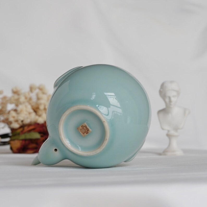 May include: A light blue ceramic teapot with a rounded body and a small spout. The teapot has a small, decorative flower emblem on the bottom. The teapot is a light, pastel blue color.