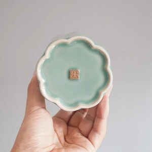 May include: A celadon-colored ceramic dish with a scalloped edge. The dish has a square, gold-colored stamp in the center. The dish is held in a person's hand.