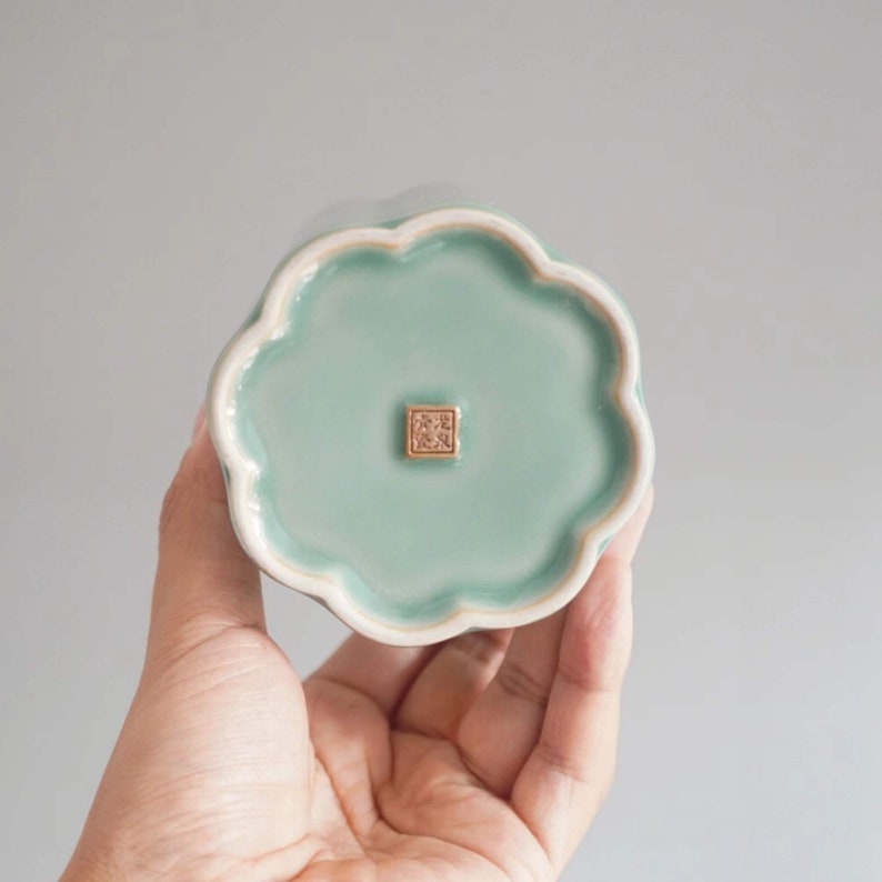 May include: A celadon-colored ceramic dish with a scalloped edge. The dish has a square, gold-colored stamp in the center. The dish is held in a person's hand.
