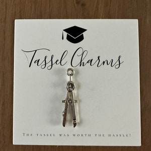 May include: A silver-toned metal drafting compass charm attached to a white display card. The card features a black graduation cap icon, the text Tassel Charms, and the phrase The tassel was worth the hassle printed in black ink.