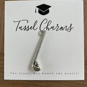 May include: A silver-toned metal golf club charm with a textured handle and a small golf ball at the base. The charm is attached to a white display card featuring a black graduation cap icon, the text Tassel Charms, and the phrase THE TASSEL WAS WORTH THE HASSLE!