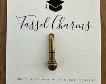 May include: A bronze-coloured microphone charm attached to a white display card. The card features a black graduation cap icon, the text "Tassel Charms" in a script font, and the text "THE TASSEL WAS WORTH THE HASSLE!" in a sans-serif font.