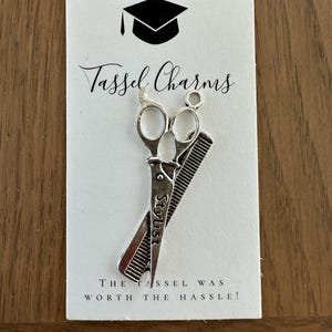 Może przedstawiać: Srebrzysty metalowy charms w kształcie nożyczek i grzebienia, z wygrawerowanym na nożyczkach słowem Stylist. Charms jest przymocowany do białej karty z ikoną czapki absolwenta, napisem Tassel Charms oraz tekstem The Tassel Was Worth The Hassle!