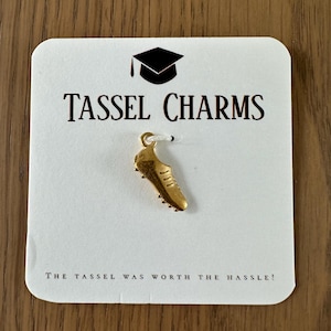 May include: A gold-colored shoe charm is displayed on a white card with the words "Tassel Charms" and a graduation cap graphic. The text "The Tassel Was Worth The Hassle!" is printed at the bottom. The charm is a miniature sneaker.