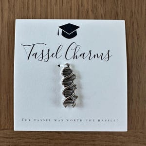 May include: A silver-toned DNA double helix charm attached to a white display card. The card features a black graduation cap icon, the text "Tassel Charms" in a script font, and the text "THE TASSEL WAS WORTH THE HASSLE!" at the bottom.