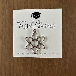 May include: A silver tassel charm shaped like an atom with the text "Tassel Charms" and "The tassel was worth the hassle!" on a white card.