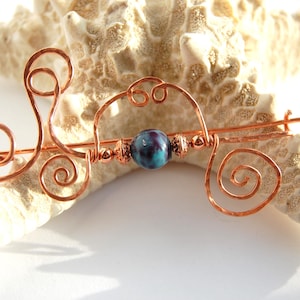 May include: A copper hair slide with a swirling design and a central blue and purple bead. The slide is made of copper wire and has a decorative, handcrafted appearance. The slide is approximately 13 cm long.