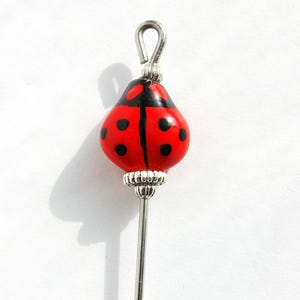 Puede incluir: Alfiler decorativo con tema de mariquita con una cuenta pintada de rojo y negro. La cuenta tiene lunares negros y una raya negra en el centro. El alfiler tiene un vástago de metal plateado y un lazo en la parte superior.