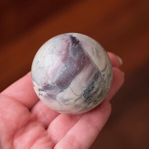 Puede incluir: Una esfera de piedra pulida y lisa con un patrón jaspeado de vetas en color blanco crema, ciruela intenso y gris carbón oscuro. El objeto esférico tiene un acabado brillante y muestra variaciones minerales naturales en su superficie.