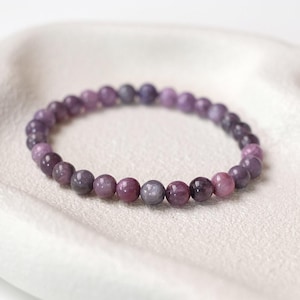 May include: A bracelet made of round, polished gemstones in shades of purple, lavender, and mauve. The bracelet is strung on an elastic band. The gemstones are approximately 0.8 cm in diameter. It rests on a white surface.