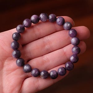 May include: A circular stretch bracelet composed of smooth, polished, spherical beads in varying shades of deep purple and lavender. The beads feature natural marbling and subtle color variations, creating a consistent, rounded design.