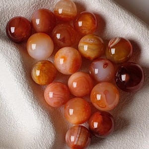 May include: A selection of polished, spherical gemstones in shades of orange, red, and brown. The stones have a glossy finish and are arranged on a white, textured surface. The gemstones vary in colour and pattern.