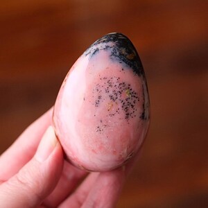 May include: A smooth, egg-shaped gemstone with a pink hue. The top of the stone features a black and white pattern, with tiny black speckles scattered across the pink surface. The stone is held in a hand and appears polished.