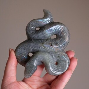 Op de afbeelding: Een gebeeldhouwde grijze edelsteen slangenbeeld. De slang is opgerold met een getextureerd, kruisgehatcherd patroon. De sculptuur heeft iriserende kleurflitsen. De slang heeft twee open oog gaten en een gebogen kop.