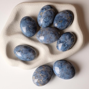 Puede incluir: Una colección de piedras azules de forma ovalada con vetas doradas, dispuestas en una bandeja blanca texturizada. Las piedras tienen un acabado pulido y brillante, que refleja la luz. La disposición está sobre una superficie blanca, creando una estética limpia y minimalista. Probablemente son piedras decorativas.