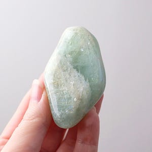 May include: A pale green gemstone with a smooth, translucent appearance. The stone has a rounded, irregular shape and a subtle, shimmering surface. The colour is a soft, muted green with areas of white and hints of iridescence. The gemstone is held in a hand.