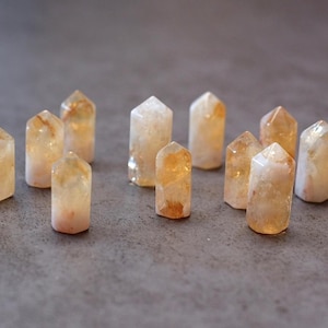 May include: A collection of small, polished citrine crystal points. The crystals are a translucent yellow-orange color, with variations in tone. They are arranged on a gray surface, each with a pointed top and a rectangular base. The crystals are a natural, earthy product.