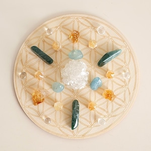 Op de afbeelding: Een cirkelvormig, lichtgekleurd houten bord met een gegraveerd geometrisch levensbloempatroon. Op het oppervlak zijn diverse kristallen gerangschikt, waaronder een centrale witte kwartsgroep, blauwe gepolijste stenen, gele citrien en helder kwarts.