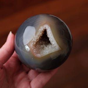 May include: A polished, spherical agate stone featuring a smooth, translucent grey surface with subtle banding. A triangular geode opening is centred on the sphere, revealing a hollow interior lined with small, sparkling, light-coloured crystals.