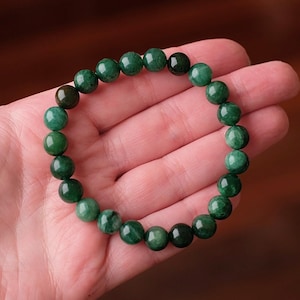 May include: A circular stretch bracelet composed of numerous smooth, round beads in varying shades of deep forest green and emerald. The beads exhibit natural colour variations and subtle internal textures, strung together to form a continuous loop.