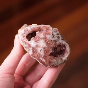 Può includere: Un esemplare minerale grezzo e irregolare caratterizzato da un interno cristallino color rosa polvere e rosa pallido con due distinte cavità vuote. La superficie esterna è ruvida, terrosa e color marrone chiaro, in contrasto con le scintillanti formazioni minerali rosa traslucide presenti all'interno della struttura a geode aperta.