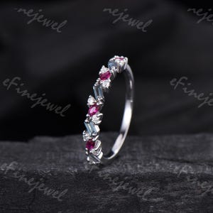 May include: A silver ring with a row of alternating gemstones. The ring features small, round white diamonds, rectangular light blue stones, and oval-shaped red stones. The ring is set against a dark background.