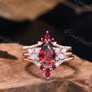 May include: A rose gold ring set featuring a large, pear-shaped, deep red gemstone as the centerpiece. The ring is adorned with smaller red and white gemstones, and opal-like stones. The ring is set on a wooden surface.
