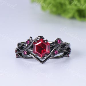 May include: A black metal ring featuring a central hexagonal red gemstone surrounded by smaller round red stones. The band is designed with a vine-like motif, incorporating sculpted leaves and branches that frame the central setting.