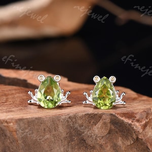 May include: A pair of sterling silver frog-shaped stud earrings. Each earring features a pear-cut, light green gemstone body with small, round, yellow-green accents for eyes. The earrings are set on a wooden surface.