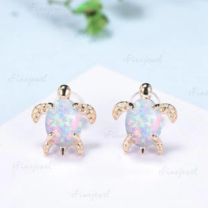 May include: A pair of gold-toned stud earrings shaped like turtles. Each turtle features a large, oval-shaped iridescent opal cabochon as the shell, displaying flashes of pink, blue, and green. The head and four legs are textured with a gold finish.
