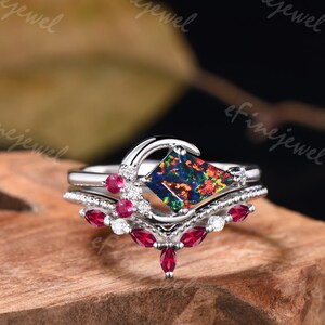May include: A silver ring set featuring a square-cut, multi-colored opal gemstone. The ring is accented with small, round red gemstones and clear crystals. The set includes a curved band with additional red gemstones.