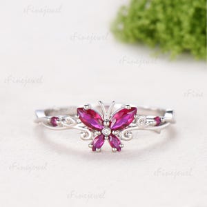 May include: A silver-toned ring featuring a central butterfly motif crafted from four marquise-cut vibrant pink stones. The butterfly body is accented with a small clear round stone, and the band displays delicate vine-like metalwork details.