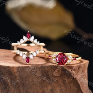 May include: Two gold rings with red gemstones and white diamonds. The top ring is a delicate, curved band with a marquise-shaped red gemstone in the center. The bottom ring has a round red gemstone surrounded by smaller white diamonds.
