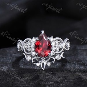May include: A pear-shaped red gemstone ring with a silver band and intricate detailing. The ring features a delicate design with smaller gemstones surrounding the main stone.