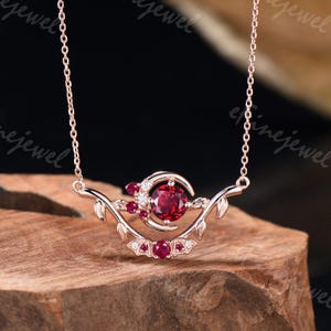 May include: Rose gold necklace featuring a central, large, round, red gemstone, surrounded by smaller red gems and clear accent stones. The design includes a crescent shape and leaf-like details, suspended from a delicate chain.