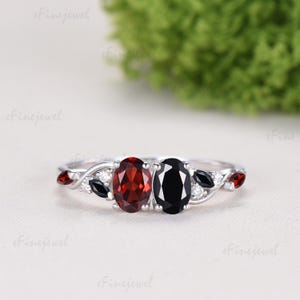 May include: A silver ring featuring two oval gemstones, one red and one black, set side by side. The band has a delicate, vine-like design with small red and black accents and tiny white stones.