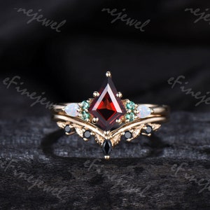 May include: A gold ring set with a large red gemstone, surrounded by smaller green and white gemstones. The ring has a delicate, ornate design with black accents.
