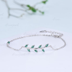 May include: A silver-toned chain bracelet featuring a central leaf-like branch motif adorned with six marquise-cut green stones. The delicate metal chain includes a lobster claw clasp and an adjustable extender link section for a secure fit.