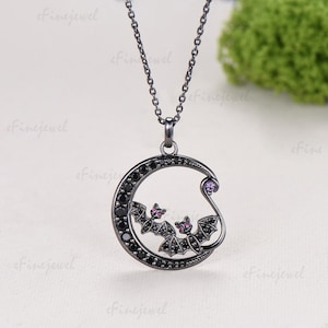May include: A black metal pendant necklace featuring a crescent moon shape encrusted with small black stones. Two small bat figures with pink stone accents are positioned within the moon, and a single purple stone sits at the top curve.