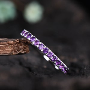 May include: A delicate silver ring featuring a continuous row of small, vibrant purple gemstones. The ring is set against a dark, textured background, highlighting the sparkle of the gemstones. The ring is a piece of jewelry.