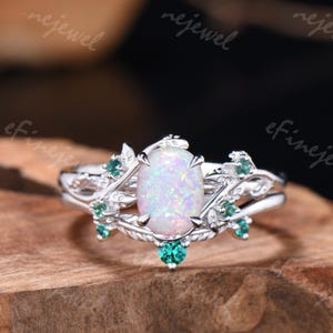 May include: A silver ring featuring an oval opal center stone, surrounded by leaf and vine detailing. Accented with small, round, emerald-green gemstones. The ring is set on a wooden surface.