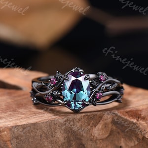May include: A black metal ring with an oval, color-changing gemstone at the center. The ring is adorned with small, round, pink gemstones and leaf-like details. The ring is set on a wooden surface.