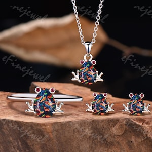 May include: A jewelry set featuring a frog design. The set includes a pendant necklace, a ring, and a pair of earrings. Each piece has a silver-colored frog with a black, blue, and orange opal body and red eyes. The jewelry is displayed on a wooden surface.