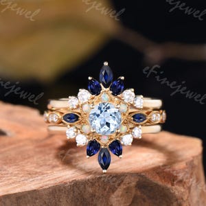 May include: A three-piece engagement ring set featuring a round aquamarine center stone surrounded by smaller white and blue gemstones. The ring is set in yellow gold.