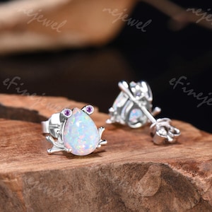 Pode incluir: Um par de brincos de prata. Cada brinco apresenta uma pedra de opala iridescente em forma de gota com duas pequenas pedras roxas. Os brincos têm um design de rã com detalhes prateados. Estão sobre uma superfície de madeira.