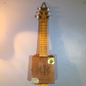 May include: A wooden cigar box guitar with a natural wood finish. The guitar has six strings and is mounted on a wall. The cigar box has a label that says "Imported", "Ashton", and "Dominican Republic".