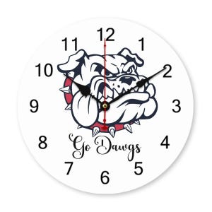 May include: A round wall clock with a white face, black numerals, and black hour and minute hands. The center features a graphic of a snarling bulldog wearing a red spiked collar. Below the bulldog, the text Go Dawgs is printed in black.